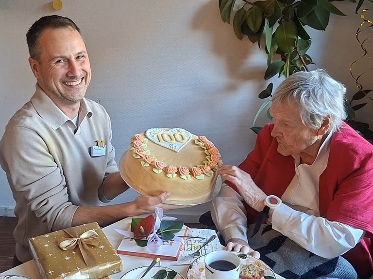 Martin Braungardt überreicht an einem Kaffetisch sitzend eine Torte mit einer verzierten 100 Martin Braungardt überreicht an einem Kaffetisch sitzend eine Torte mit einer verzierten 100