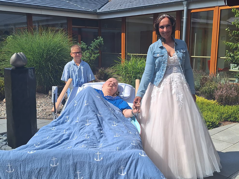 Gästin des Albertinen Hospiz Norderstedt hält im sonnigen Innenhof die Hand ihrer Tochter im Hochzeitskleid; ihr Enkel steht hinter dem Bett. Gästin des Albertinen Hospiz Norderstedt hält im sonnigen Innenhof die Hand ihrer Tochter im Hochzeitskleid; ihr Enkel steht hinter dem Bett.